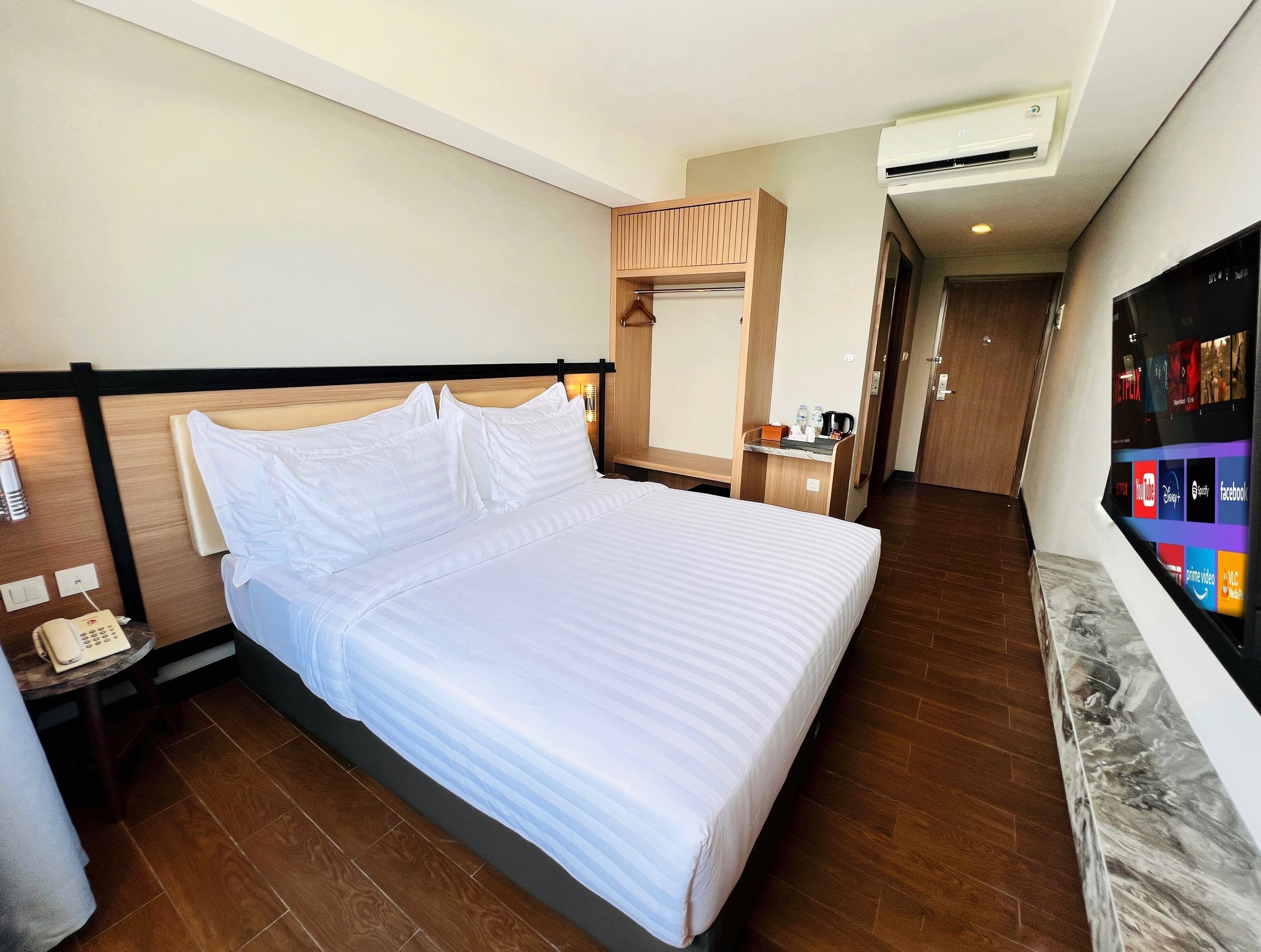 A hotel room with a large bed, a bedside table with a phone, a small table with a kettle, and a wall-mounted TV displaying various channels.