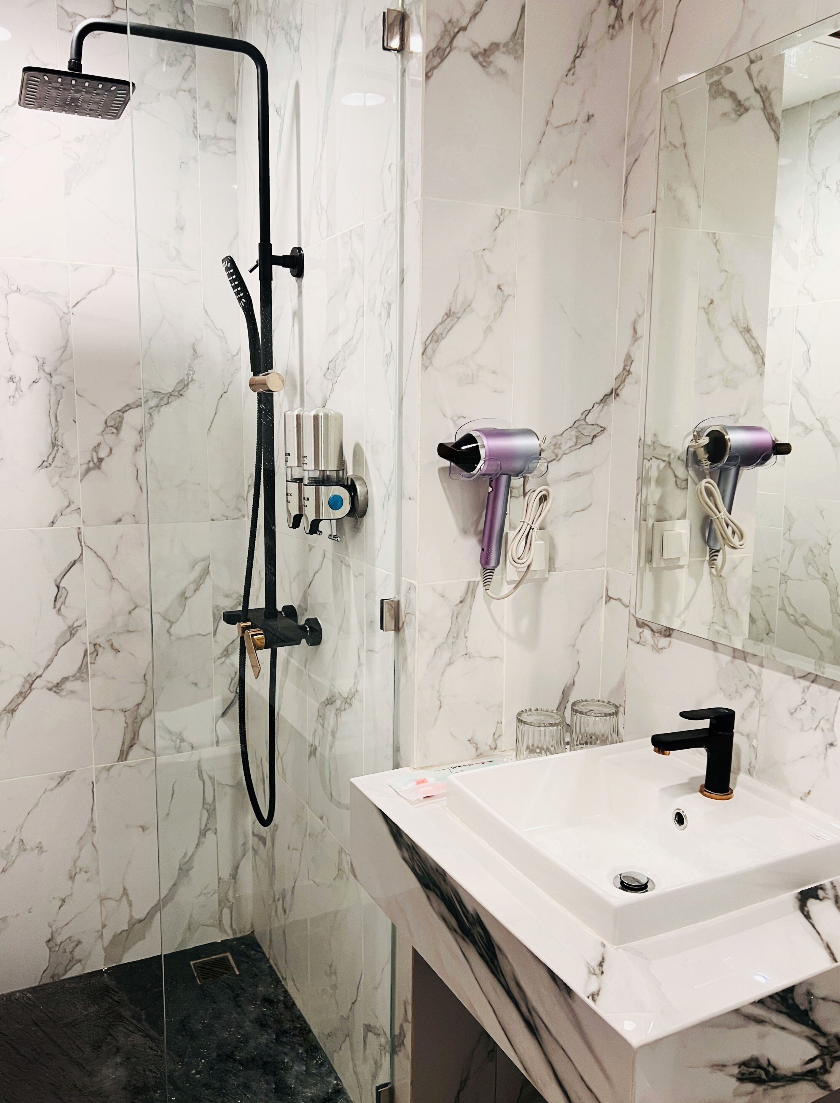Un baño con pared de mármol, ducha, lavabo y dos secadores de pelo fijados a la pared.