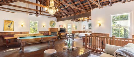 A spacious living room with a pool table, comfortable seating, a fireplace, and a large window offering a view of the outdoors.