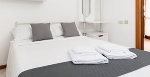 Una cama bien hecha con sábanas blancas y almohadas grises, una mesita de noche con una lámpara y una toalla doblada sobre la cama.
