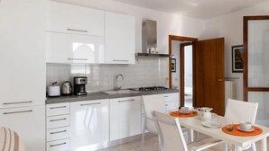 Una cocina moderna con armarios blancos, estufa a gas y cafetera. Hay un comedor con una mesa y sillas blancas.
