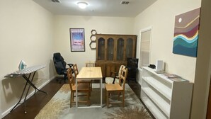 A dining area with a wooden table and chairs, a small ironing board, and a cabinet with shelves. - Cjay Homes (Jersey City)