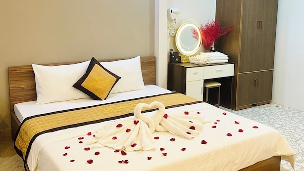 A neatly made bed with a heart-shaped towel arrangement and rose petals, a bedside table with a mirror and decorative items, and a wardrobe in a hotel room.
