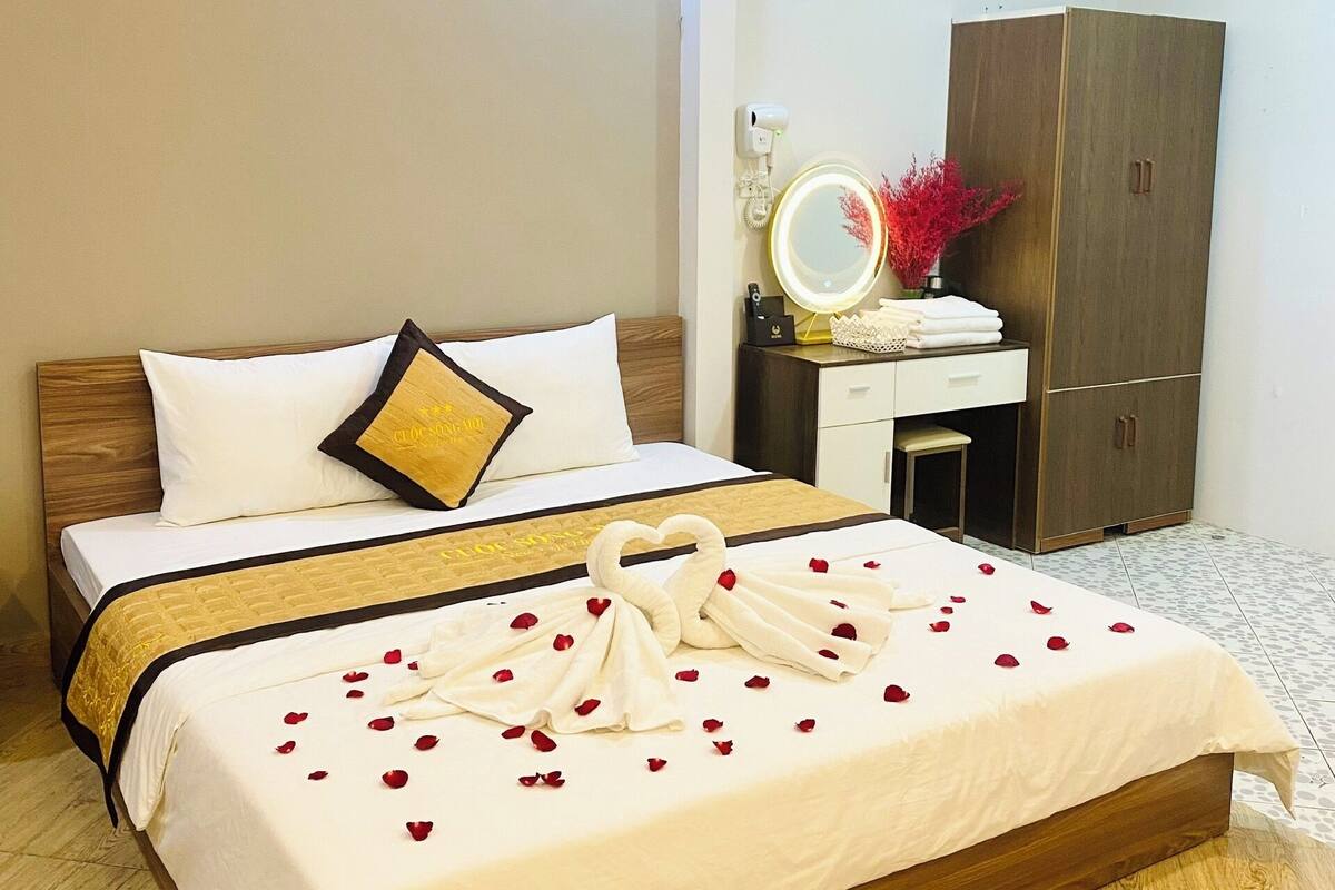 A neatly made bed with a heart-shaped towel arrangement and rose petals, a bedside table with a mirror and decorative items, and a wardrobe in a hotel room.