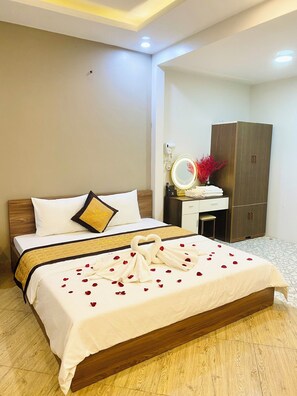 A neatly made bed with a heart-shaped towel arrangement and rose petals, a bedside table with a mirror and decorative items, and a wardrobe in a hotel room. - Newlife Hotel LB - by BAY LUXURY (Hanoi)