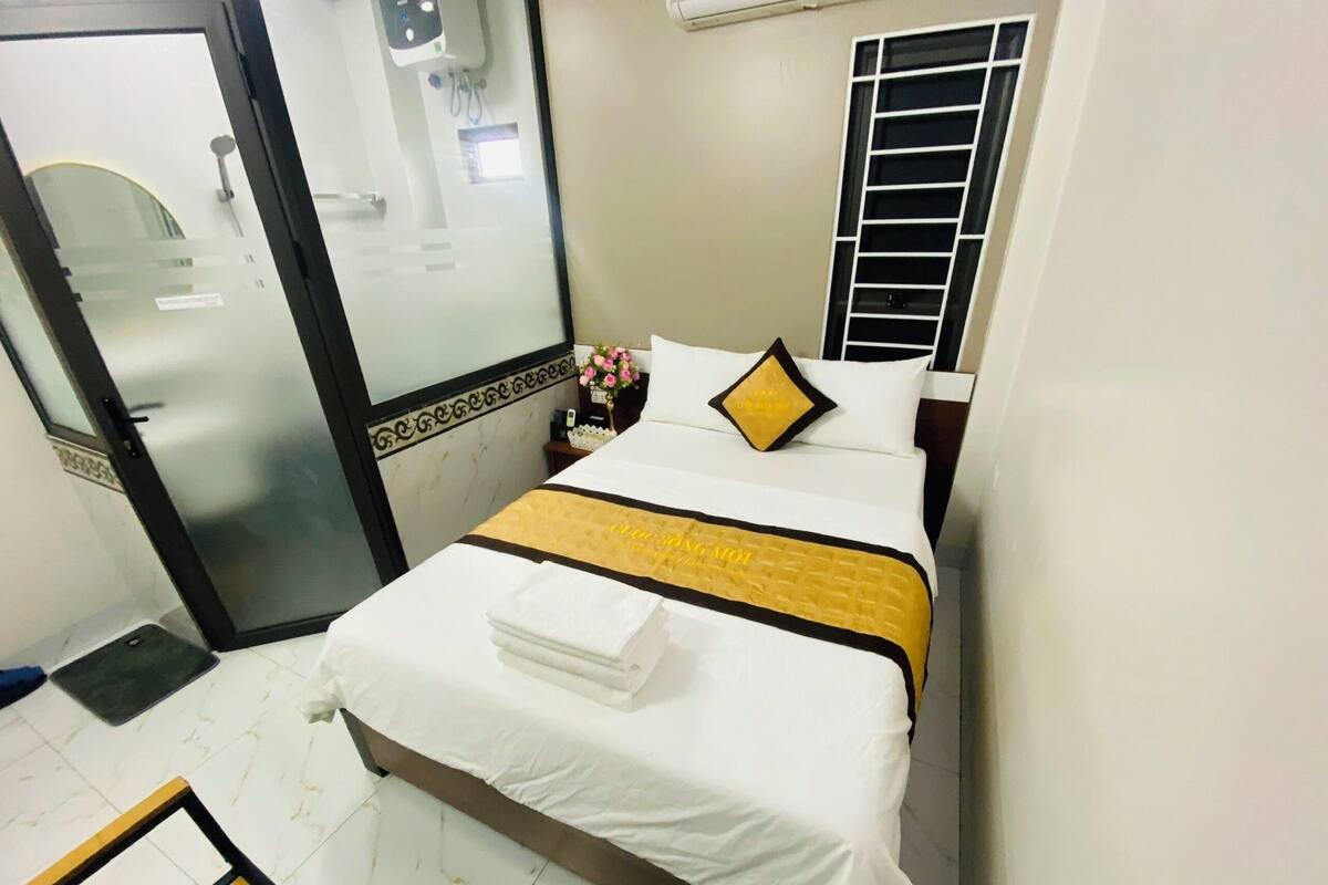 A hotel room with a bed, a bedside table, a mirror, a window with blinds, and a door.