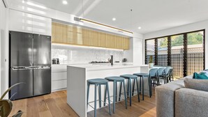 A modern kitchen with a stainless steel refrigerator, wooden cabinets, and a marble countertop. There are four blue bar stools and a grey sofa in the living area. - Salt Life Byron. Wifi, Parking & Pool in Paradise (Byron Bay)