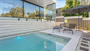 A modern outdoor pool area with a glass wall, lounge chairs, and a wooden deck. - Salt Life Byron. Wifi, Parking & Pool in Paradise (Byron Bay)