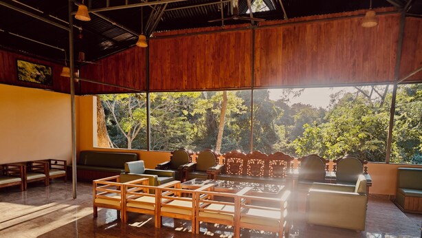 A spacious room with a large window offering a view of lush greenery. There are several wooden chairs and a sofa arranged for seating. - KAZIRANGA ECO VILLAGE RESORT (Kaliabar)