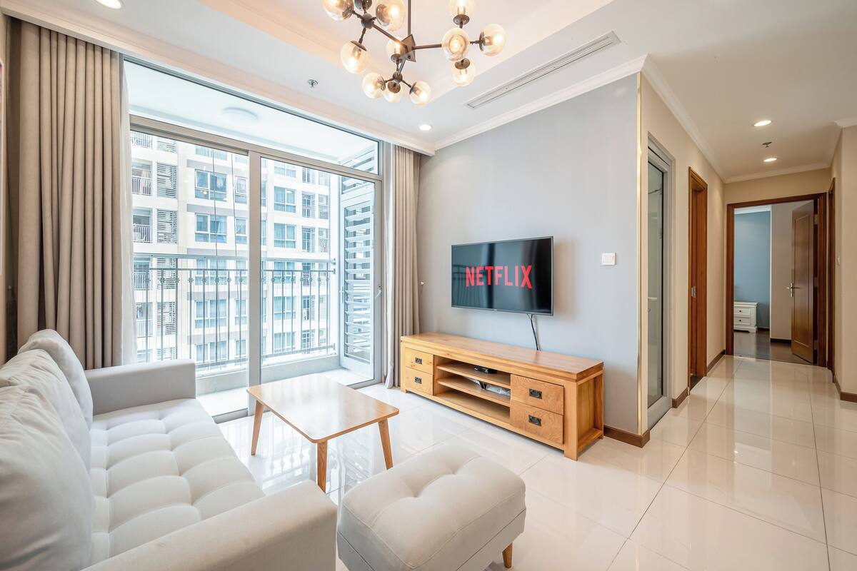 A modern living room with a large window, a sofa, a coffee table, a wooden TV stand, and a flat-screen TV displaying "NETFLIX".