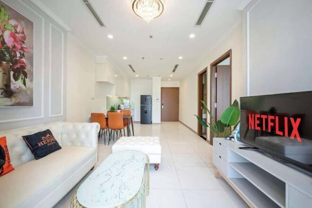 A modern living room with a white sofa, a coffee table, a TV with 'NETFLIX' sign, and a dining area in the background.