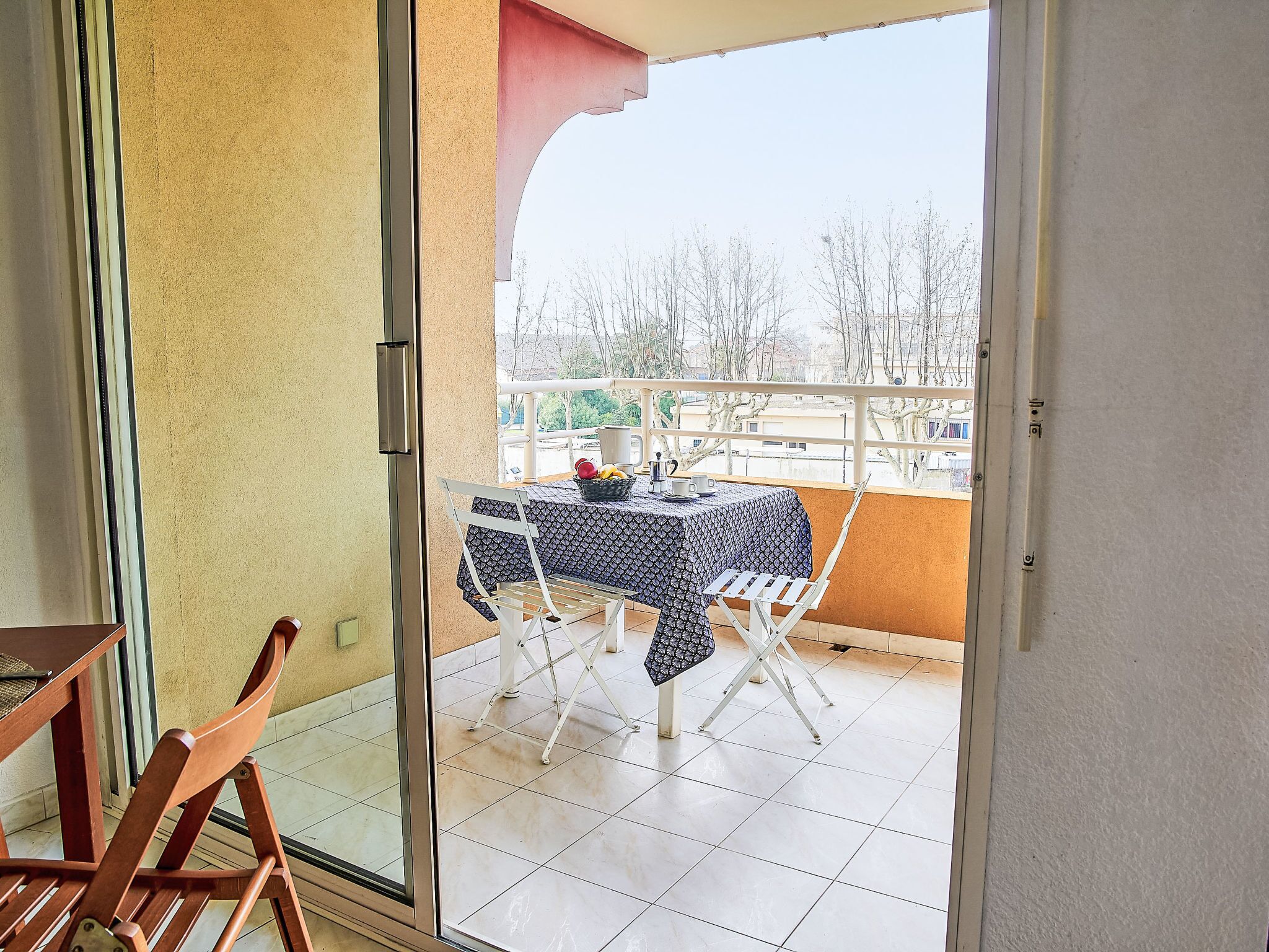 A balcony with a table and chairs, a view of trees and buildings, and a door leading to the interior.