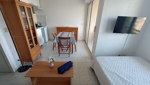 A compact room with a bed, a small table with a vase, a wooden cabinet, a dining table with chairs, and a TV mounted on the wall. - Les Rives Latines by Interhome (Fréjus)
