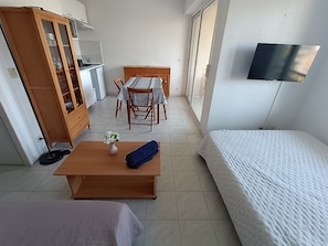 Une pièce de taille modeste comprenant un lit, une petite table avec un vase, un meuble en bois, une table à manger avec des chaises et un téléviseur fixé au mur.