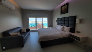 Un dormitorio con una cama, un sofá de color oscuro, una mesita de noche y un ventanal con vistas al mar.