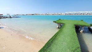 Una playa con un muelle de hierba que se adentra en el agua, un cielo azul despejado y una hilera de edificios blancos al fondo.