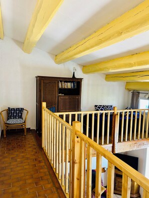 Una escalera de madera con barandilla que conduce a un balcón en el segundo piso. Se ve una estantería de madera y una silla con estampado en el primer piso.