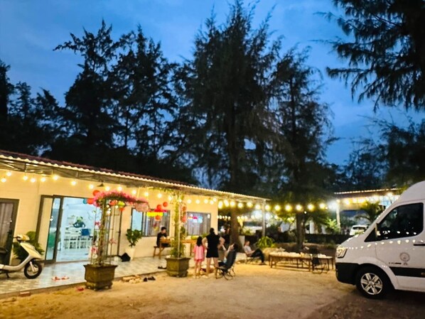 Un área de estar al aire libre con mesas y sillas, rodeada de árboles y un edificio con luces colgantes.