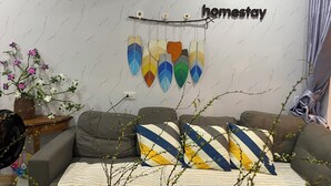 Een woonkamer met een bank, decoratieve kussens, een aan de muur bevestigd bordje met de tekst 'RUBY HOUSE homestay' en een wanddecoratie van kleurrijke vogelveren.