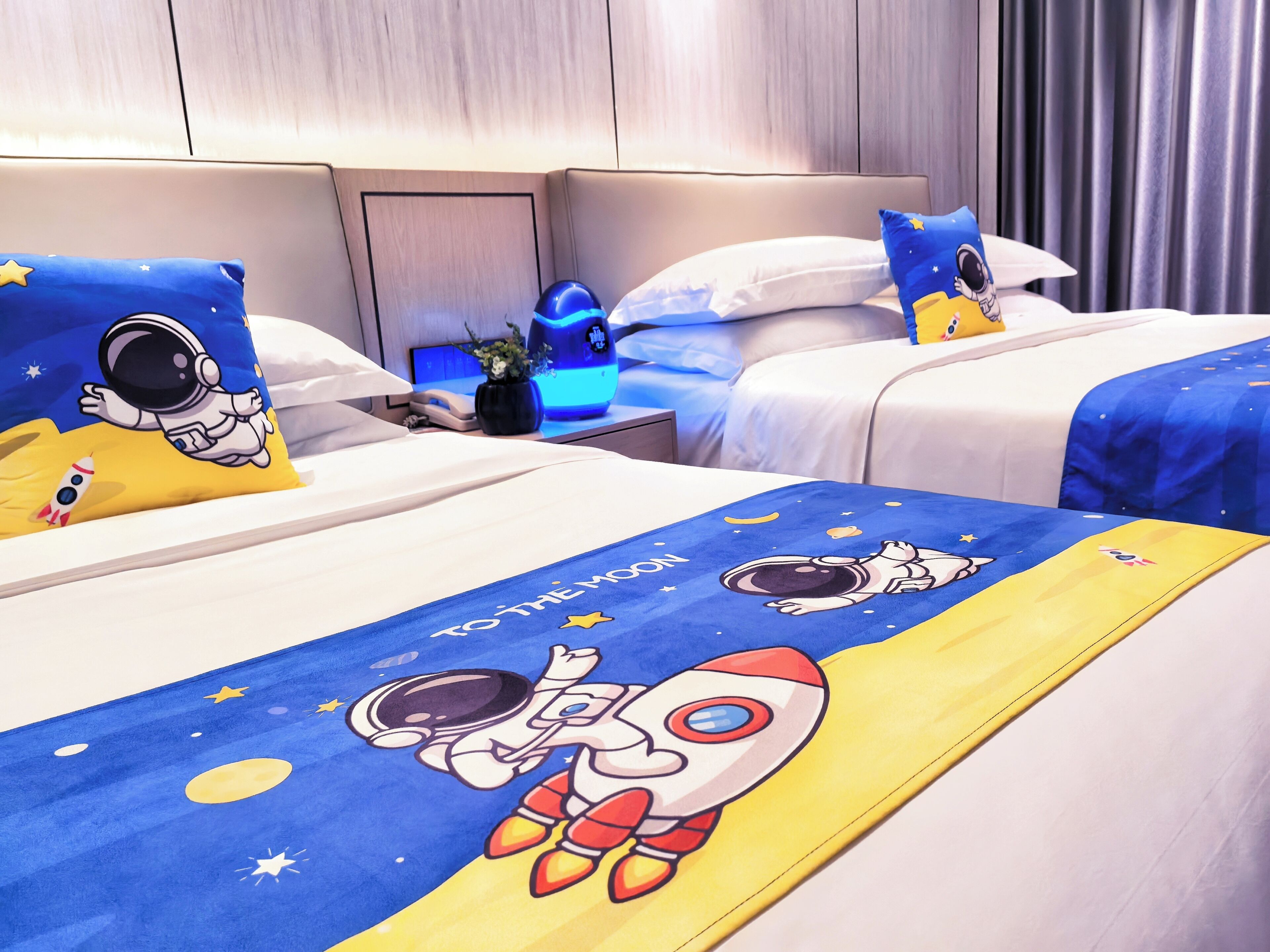 A hotel room with two beds, each with astronaut-themed bedding and pillows. There is a bedside table with a lamp and a small plant.