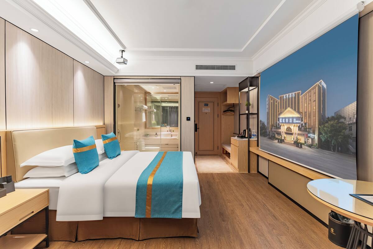 A hotel room with a large bed, a desk, a TV, and a painting of a building.