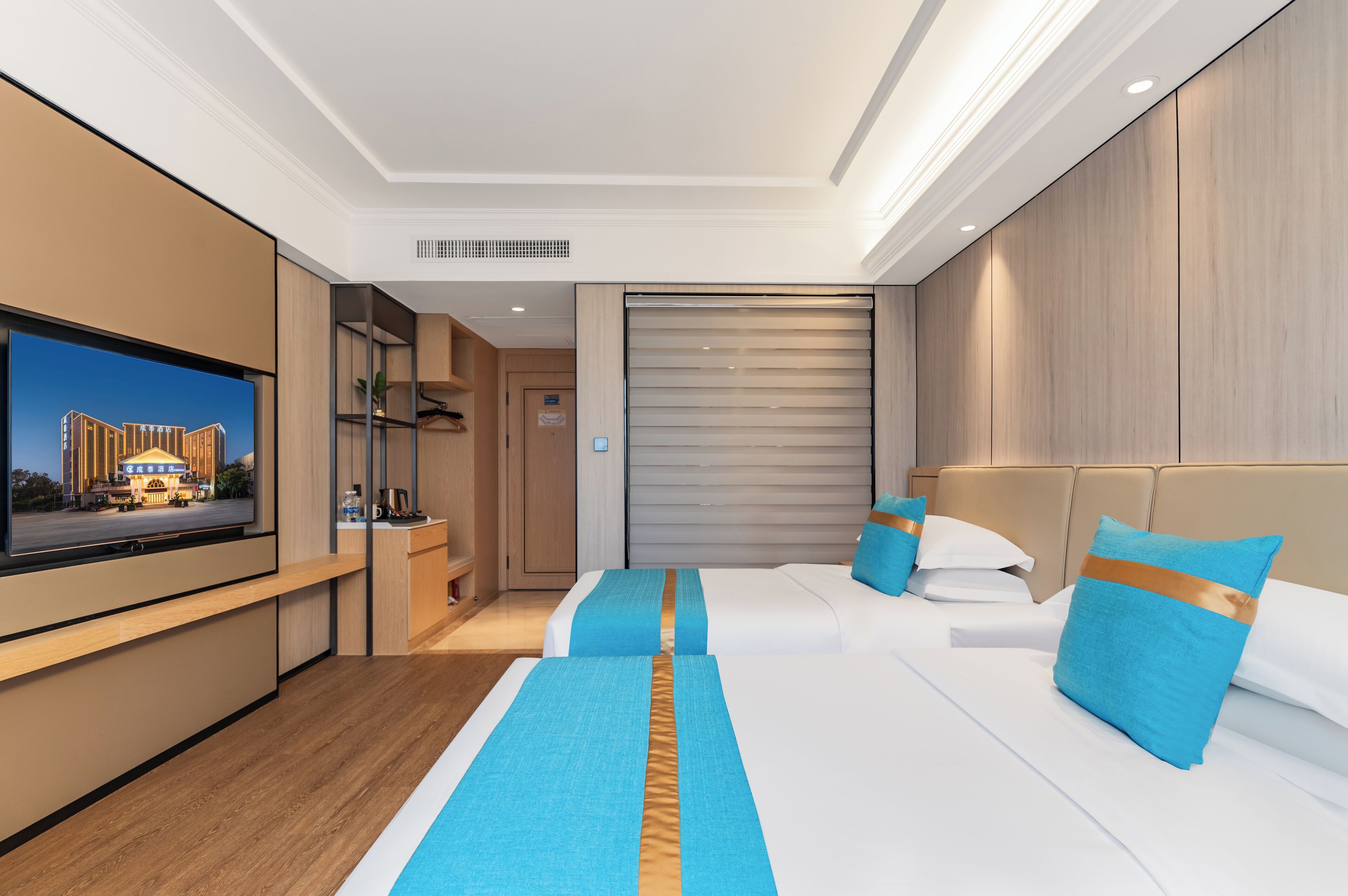 A hotel room with a large flat-screen TV, two beds with blue and gold accents, a wooden wardrobe, and a sliding door closet.