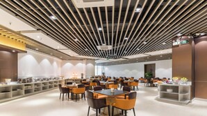 Un comedor moderno con varias mesas y sillas, una barra larga con exhibición de alimentos y un techo con luminarias lineales.