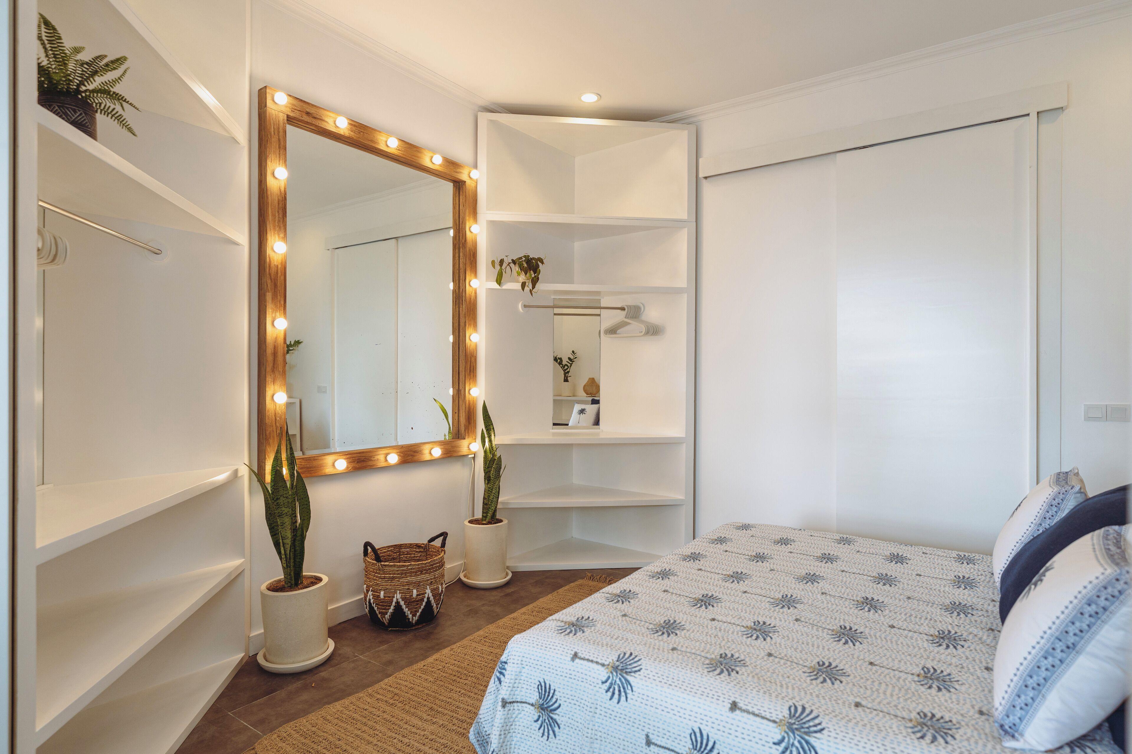 Un dormitorio con un espejo grande iluminado, una cama con ropa de cama estampada y una planta en maceta.