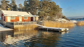 Una cabaña con un muelle que se adentra en un lago tranquilo, rodeada de árboles y un cielo despejado.