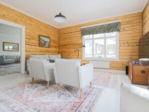 A room with wooden walls, a white sofa, a dining table with chairs, a window with curtains, and a painting on the wall. - Kapteenin huvilat by Interhome (Parainen)