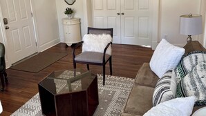 A living room with a ceiling fan, a brown sofa, a glass coffee table, a patterned rug, a white armchair, and a white door.