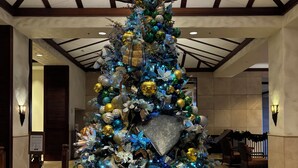 Un albero di Natale decorato con addobbi e luci, circondato da pacchetti regalo, nell'atrio di un hotel con soffitto alto e travi in legno.