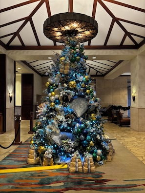 Een kerstboom versierd met versieringen en lichtjes, omringd door ingepakte cadeaus, in een hotel lobby met een hoog plafond en houten balken.