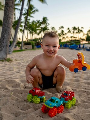 Un enfant joue avec des voitures miniatures sur une surface sablonne, avec des palmiers en arrière-plan.