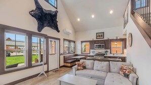 A spacious living room with a large sectional sofa, a coffee table, and a mounted animal hide. There is a kitchen area with modern appliances and a dining table. Large windows provide a view of the outdoors. - Cozy Dover Bay Pet Friendly Retreat - Sleeps 5 (Dover Bay)