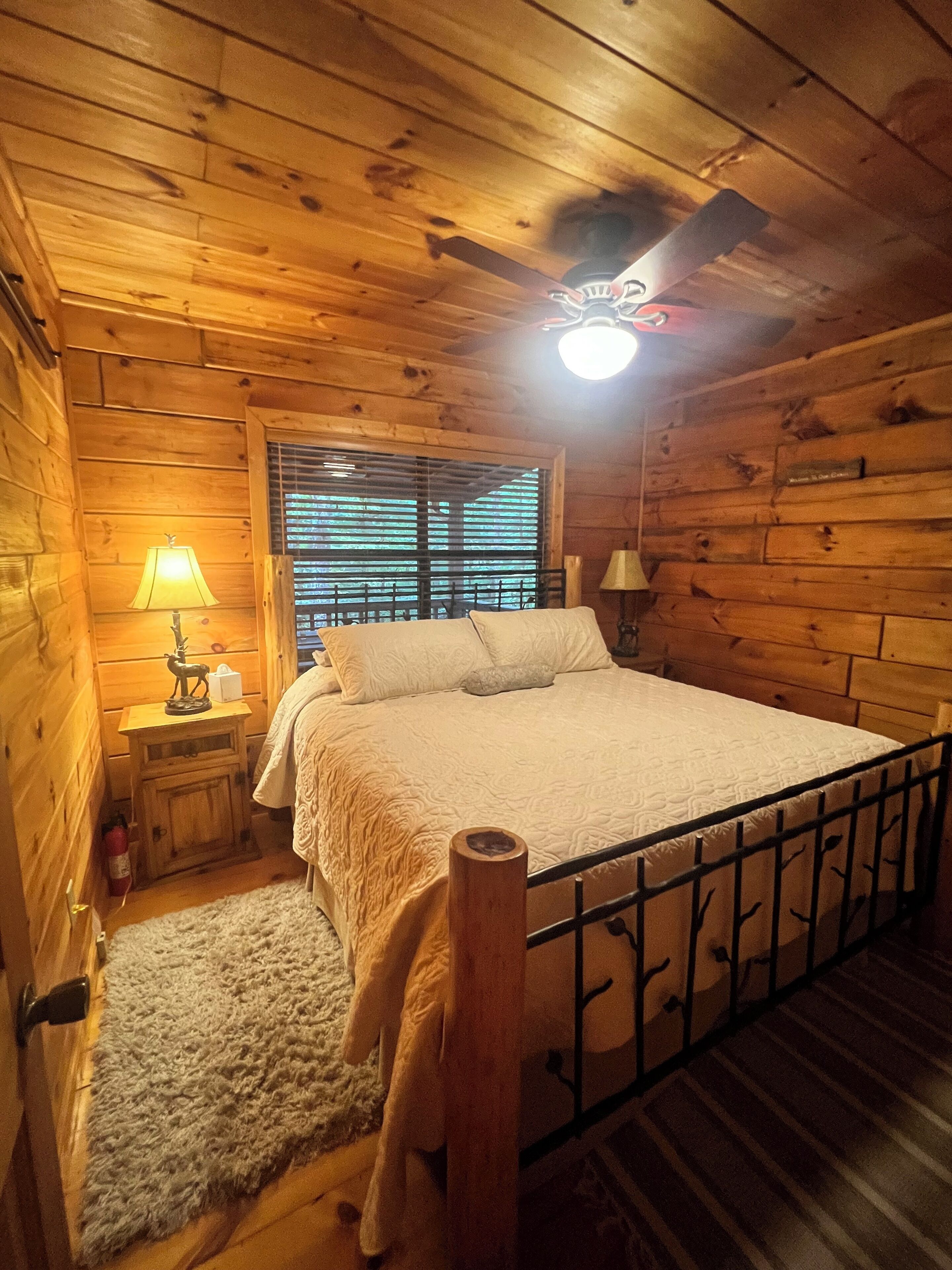 Quarto acolhedor de cabana de madeira, com uma cama, uma mesa de cabeceira com abajur, uma janela com persianas e um ventilador de teto.