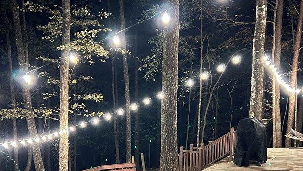 A stone deck with string lights, a wooden bench, and a covered grill area in a wooded setting. - Experience Serenity at Triple Nickel Cabin! (Blue Ridge)