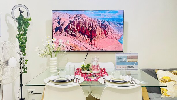 A dining area with a glass table set for two, a flat-screen TV displaying a mountain landscape, a wall-mounted fan, and a house rules sign. - MARIA CONDO 19F Cebu High-Rise (Mandaue City)