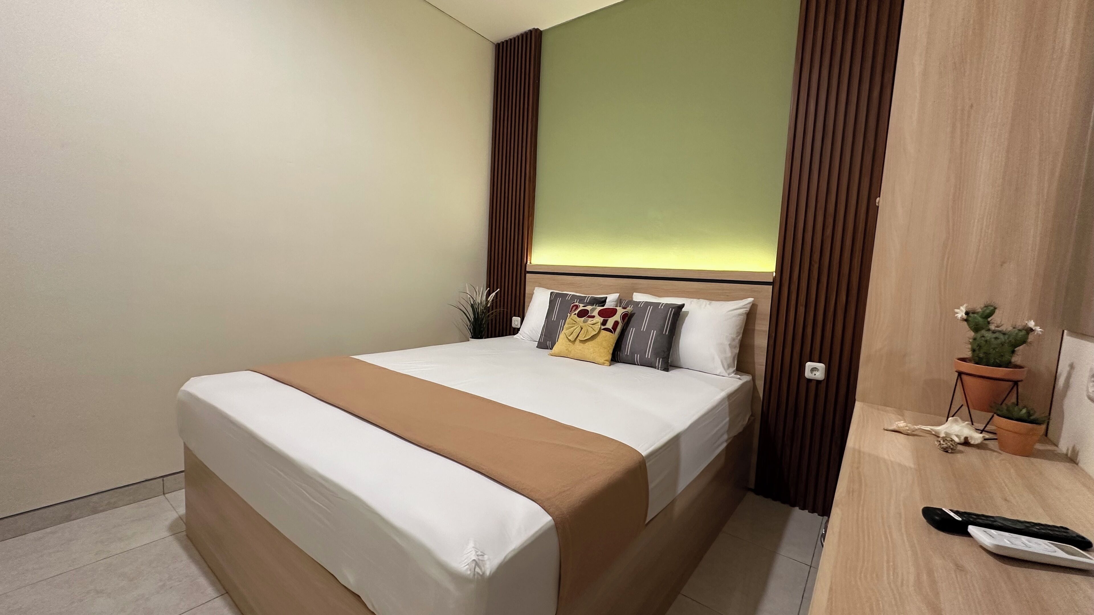 A neatly made bed with a brown bedspread and white linens, a bedside table with a remote control, and a potted cactus on the right.