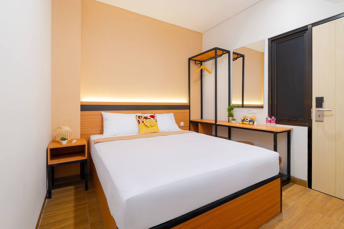 Quarto de hotel moderno com uma cama bem arrumada, mesa de cabeceira com abajur, uma pequena planta e um espelho acima da mesa.