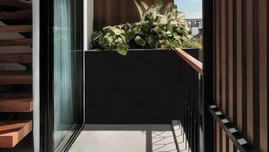 Un balcón moderno con suelo de madera, baranda negra y una jardinera con plantas verdes.