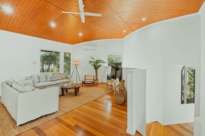 A spacious living room with a wooden ceiling, a white sofa, a coffee table, and a large window providing a view of greenery outside.