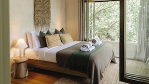Una cama bien hecha con estructura de madera, cabecero tejido y vista a la vegetación a través de un ventanal grande.