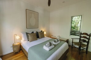 A neatly made bed with pillows, a bedside lamp, a chair, and a window with a view of greenery.