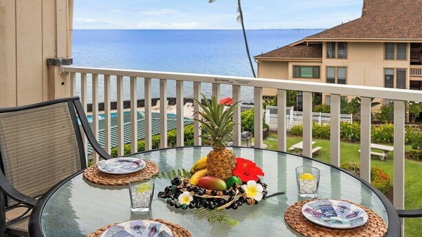Een ronde glazen tafel met een tropisch fruitstuk in het midden, twee borden en twee glazen op een balkon met uitzicht op een zwembad en de oceaan.