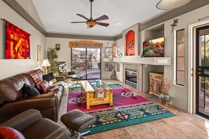 Un salon spacieux avec un canapé en cuir, une table basse en bois et un tapis coloré. On y trouve une cheminée agrémentée d’un manteau décoratif et un ventilateur de plafond.