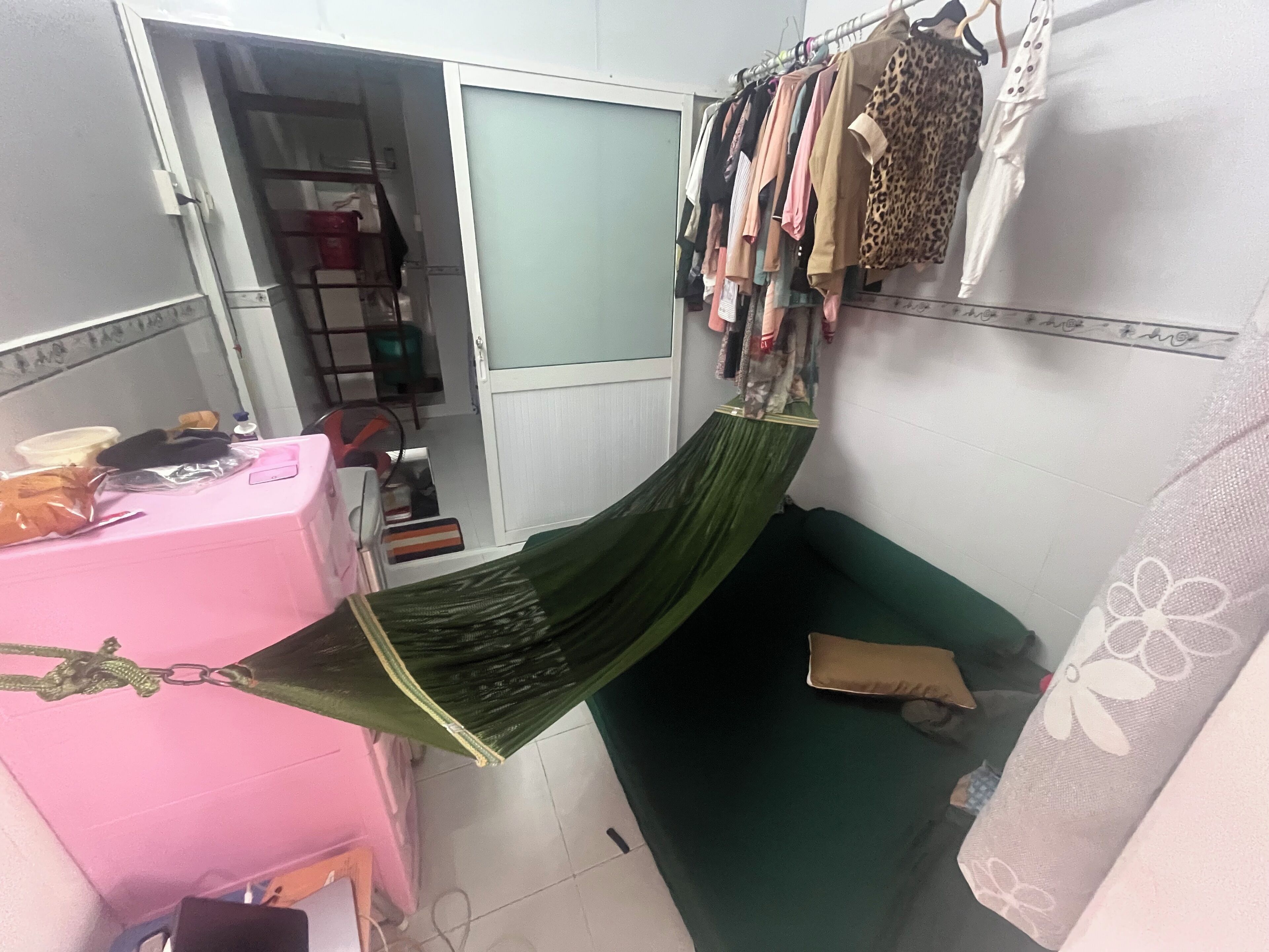 Ein kleiner, kompakter Raum mit einem grünen Bett, einer pinkfarbenen Kommode und einem Kleiderständer mit verschiedenen Kleidungsstücken.