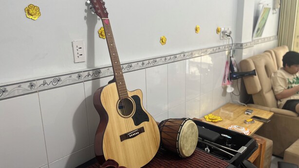 Een kamer met een gitaar die tegen de muur leunt, een trommel op een mat, en een persoon zittend in een stoel.