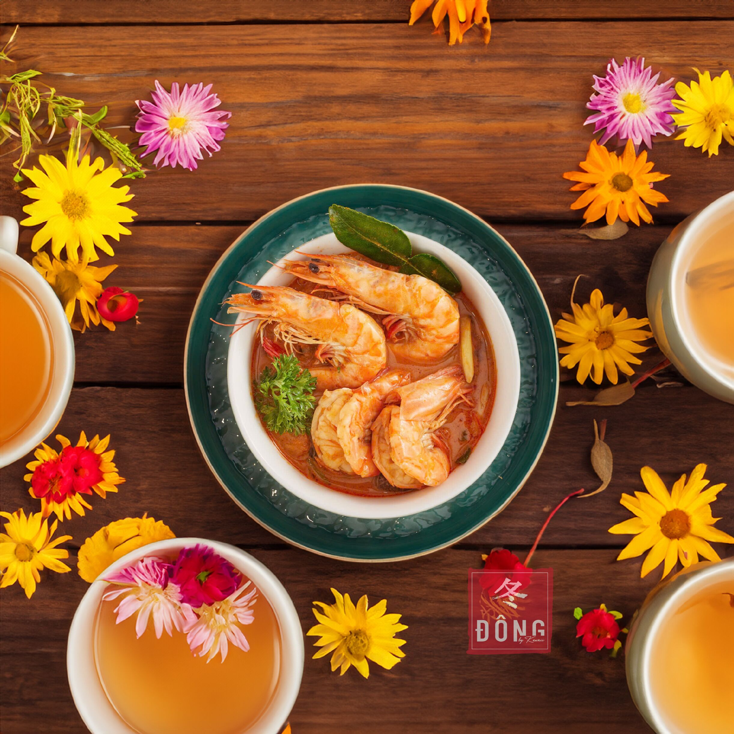 Un bol de soupe aux crevettes, entouré de fleurs et de tasses de thé, sur une table en bois.