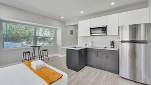 A modern hotel room with a bed, a small dining area, a kitchenette with a refrigerator, and a large window offering a view of the outdoors. - Walk to Beach Studio | Pompano Beach (Pompano Beach)